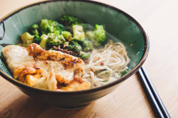 Close up of a bowl of udon noodle soup with fried egg and broccoli