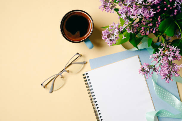 istock Flat lay home office desk. Female workspace with white paper notebook, glasses, coffee cup and lilac flowers on beige background. Top view feminine background. Fashion blogger desktop concept. 1147865340