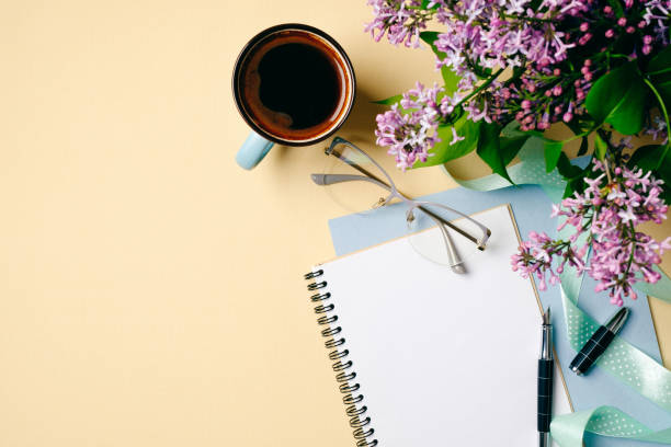 istock Flat lay home office desk. Female workspace with white paper notebook, coffee cup and lilac flowers on beige background. Top view feminine background. Fashion blogger desktop concept. 1147865342