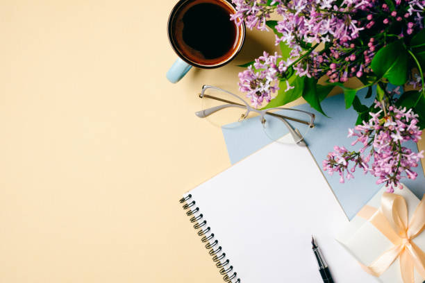 istock Flat lay home office desk. Female workspace with white paper notebook, glasses, coffee cup and lilac flowers on beige background. Top view feminine background. Fashion blogger desktop concept. 1147865346