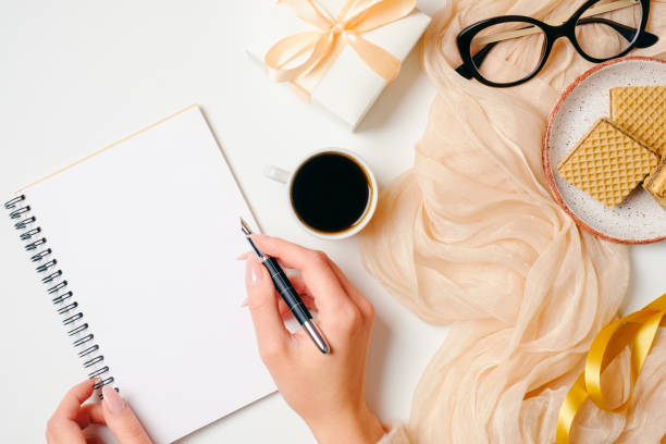 istock Women's home office desk. Workspace with female hands writing message in paper notebook and feminine golden fashion accessories on white background. Girl's diary, beauty blogger, writer work concept 1147865399