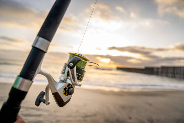 a close-up view of a fishing pole with the beach and waves at sunset near the international border wall  in playas tijuana, mexico - carretel de pesca imagens e fotografias de stock