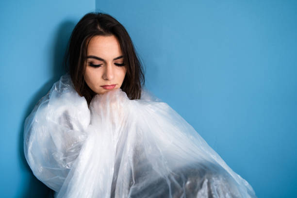 istock Young scared woman sad covered with plastic nylon cellophane sheet sitting helpless in a corner of a blue room pollution save the earth concept 1147879675