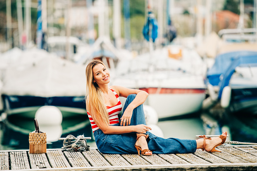 Outdoor fashion portrait of beautiful youn woman posing by the lake, wearing red and white stripe vest and denim wide leg jean