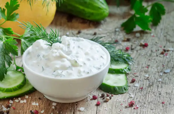 Photo of Ranch whte sauce in white bowl with cucumber, herbs and spices on old wooden table, selective focus