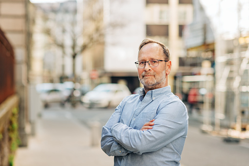 Outdoor portrait of handsome middle age man wearing long sleeve Oxford shirt, posing on city street, arms crossed