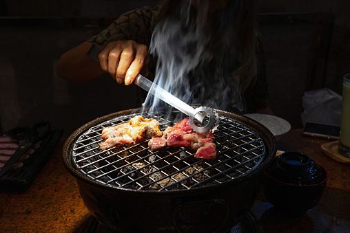 Asian female hand holding steel food tongs picking up sliced beef on charcoal grill.