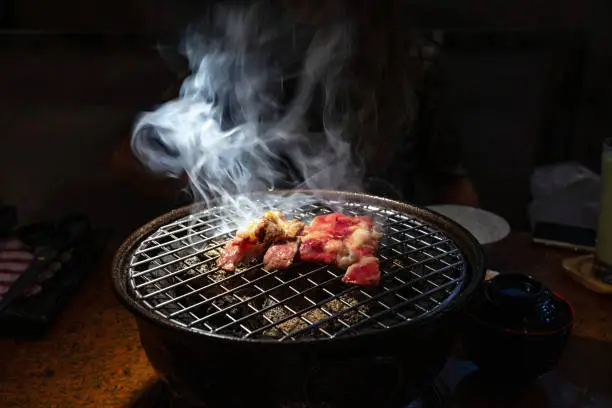 Cooked and raw beef on charcoal grill with white smoke.