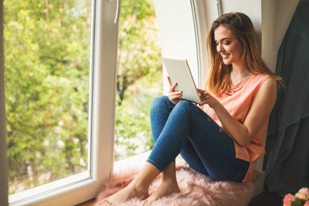 istock Woman relaxing at window sill, typing on digital tablet 1147929213