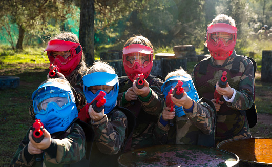 Boys and girls paintball players aiming and shooting with guns at opposing team outdoors