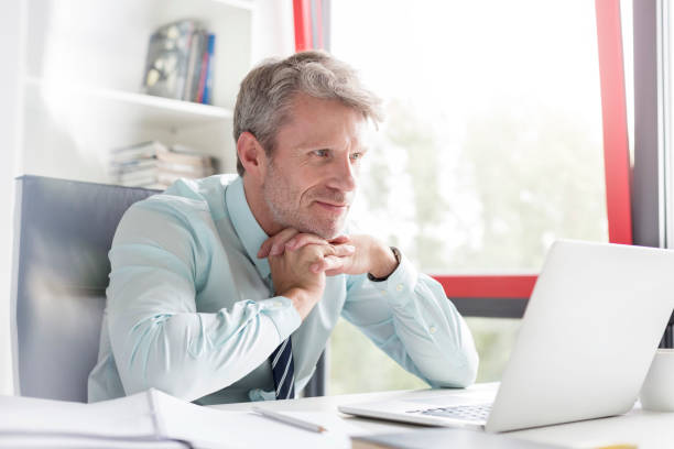 istock Smiling thoughtful businessman with hands clasped looking at laptop in office 1147936644
