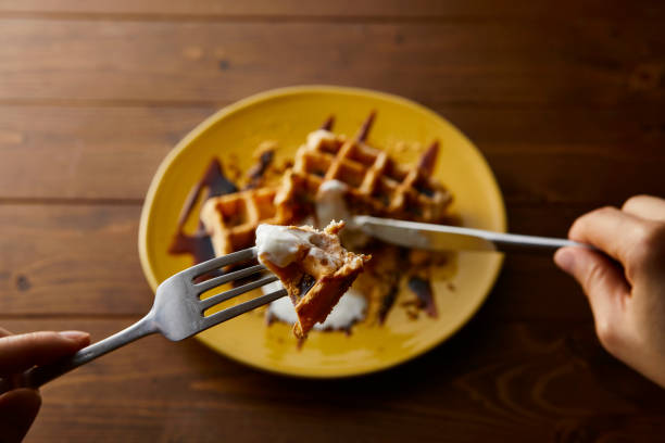 Hand eating rice flour waffle with black honey and yoghurt served on a yellow round plate.