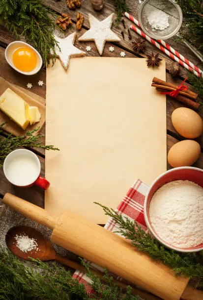 Christmas - baking cake background. Old sheet of paper with dough ingredients and decorations around on vintage planked wood table from above. Rural kitchen layout with free text space.