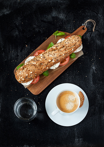 Sandwich (mozzarella cheese, tomatoes and fresh basil), coffee and water on black chalkboard background. Cafe table from above. Poster layout with free text space.