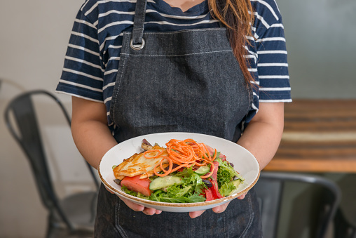 Waitress with Apron Holding a Plate of Salad with table and Chair Blur Background