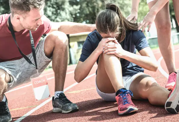 Photo of Female athlete getting injured during athletic run training - Male coach taking care on sport pupil after physical accident - Team care concept with young sporty people facing mishaps casualty
