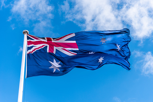 The Australian flag attached to a white flag pole, flying in the wind against a light cloud blue sky