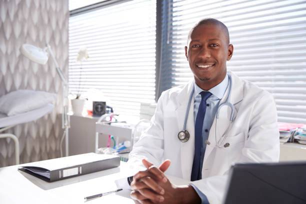 Portrait Of Smiling Male Doctor Wearing White Coat With Stethoscope Sitting Behind Desk In Office Portrait Of Smiling Male Doctor Wearing White Coat With Stethoscope Sitting Behind Desk In Office dermatology photos stock pictures, royalty-free photos & images