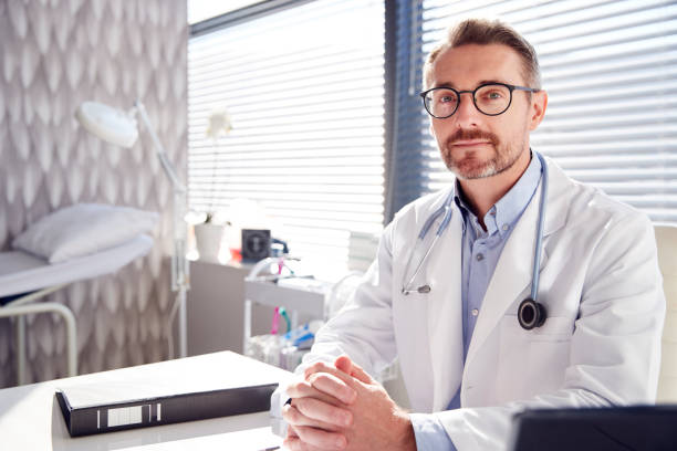 porträt von smiling male doctor wearing white coat mit stethoscope sitting behind desk im büro - doctor male doctor mature men portrait stock-fotos und bilder