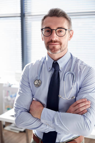 porträt des lächelnden männlichen arztes mit stethoscope standing by desk im büro - doctor male doctor mature men portrait stock-fotos und bilder