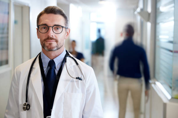 ritratto di medico maschio maturo che indossa un camice bianco con stetoscopio nel corridoio ospedaliero affollato - doctor male doctor mature men portrait foto e immagini stock