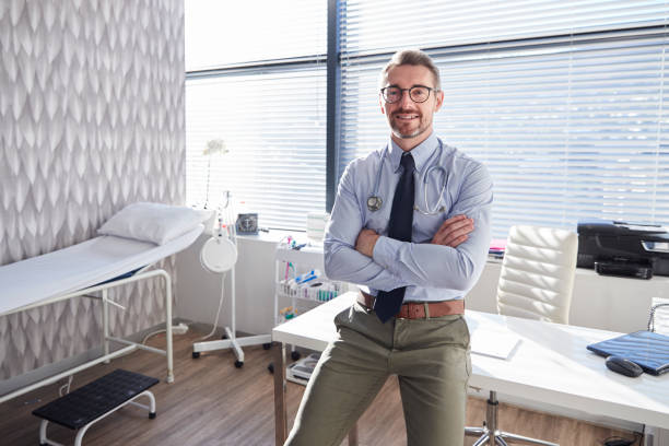 portrait of smiling mature male doctor with stethoscope standing by desk in office - medico consultorio imagens e fotografias de stock