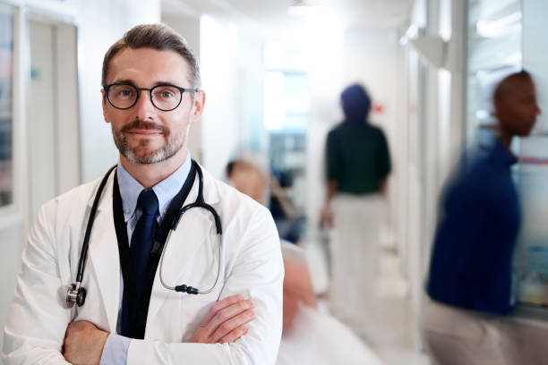 porträt der mature male doctor wearing white coat mit stethoskop in busy hospital corridor - doctor male doctor mature men portrait stock-fotos und bilder