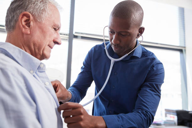 doctor listening to chest of senior male patient during medical exam in office - male african descent africa ethnic imagens e fotografias de stock
