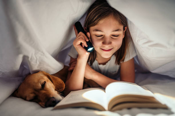 chica acostada en la cama con su perro bajo el libro de lectura de manta tarde en la noche - enjoying a novel fotografías e imágenes de stock