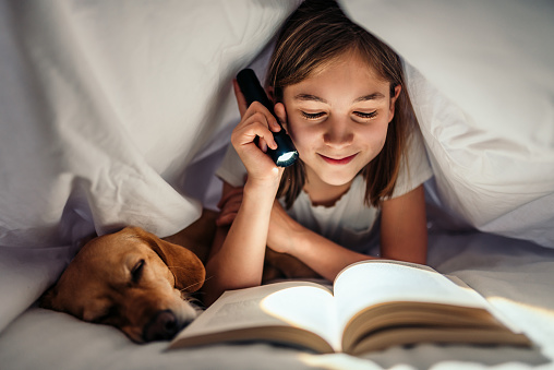 Girl lying in the bed with her dog under blanket reading book late at night