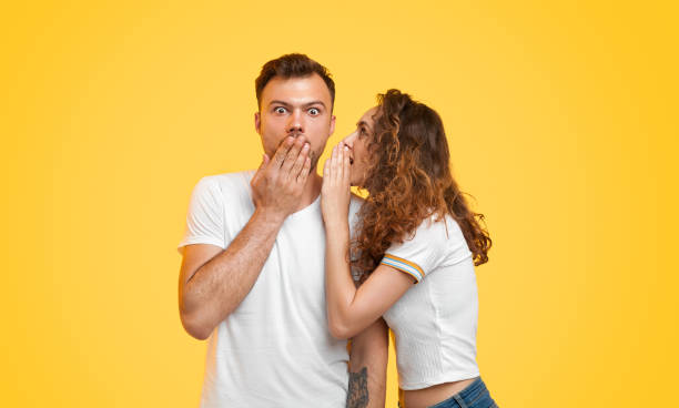 Young couple gossiping on yellow background Pretty young woman telling secret to shocked boyfriend while standing on vibrant yellow background together whispering stock pictures, royalty-free photos & images