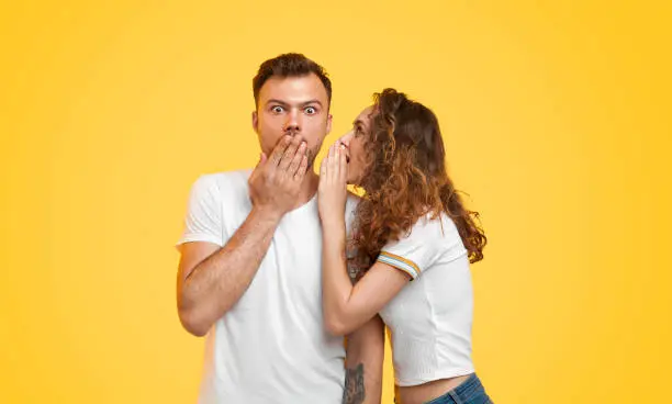 Pretty young woman telling secret to shocked boyfriend while standing on vibrant yellow background together