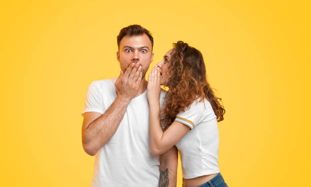 молодая пара сплетничает на желтом фоне - whispering gossip women secrecy стоковые фото и изображения
