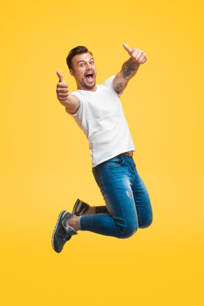 Excited man jumping and gesturing thumb up Handsome young guy in casual outfit screaming and showing thumb up gesture with both hands while looking at camera and jumping on bright yellow background jumper stock pictures, royalty-free photos & images