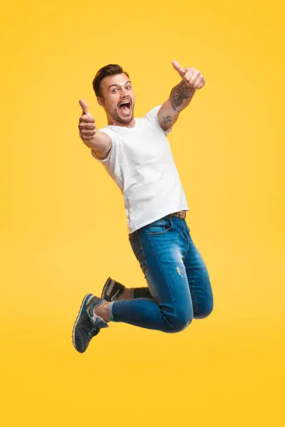Handsome young guy in casual outfit screaming and showing thumb up gesture with both hands while looking at camera and jumping on bright yellow background