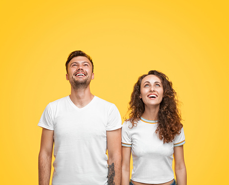 Handsome young man and beautiful woman in white T-shirts smiling and looking up while standing on yellow background