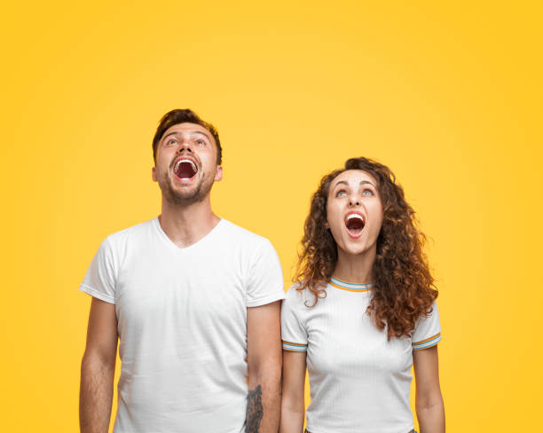Shocked couple looking up Young man and woman in white T-shirts looking up with astonished face expression while standing on bright yellow background mouth open human face shouting screaming stock pictures, royalty-free photos & images