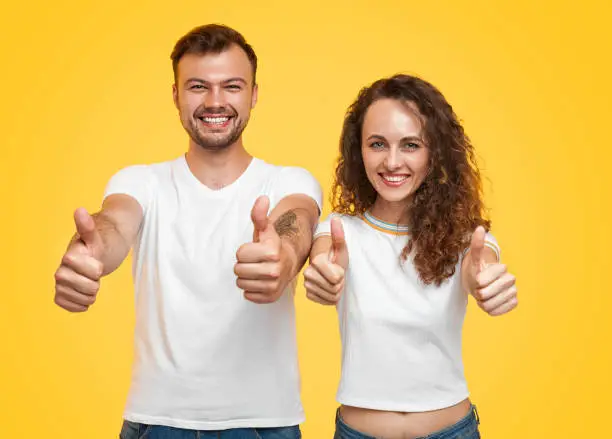 Young man and woman in white T-shirts cheerfully smiling and looking at camera while standing on bright yellow background and showing thumb-up gesture