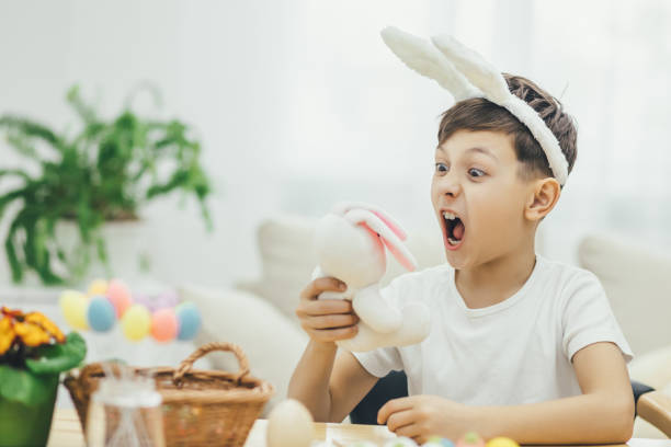 istock Happy smiling little boy in bunny ears sitting at rhe table surrounded by the easter tiny things, holding funny pink easter bunny and making grimace. 1147990819