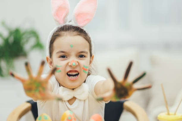 istock Little painter colouring easter eggs, showing at the camera her colored hands, showing her tongue. Rabbit ears on her head. Expressive face. 1147990947