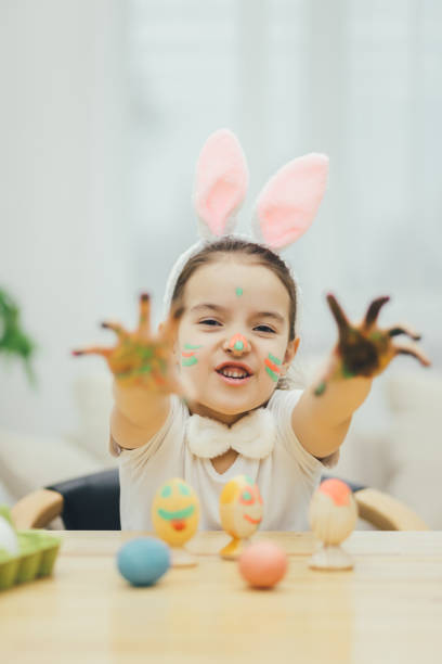 istock Little painter colouring easter eggs, showing at the camera her colored hands, showing her tongue. Rabbit ears on her head. Expressive face. 1147990962