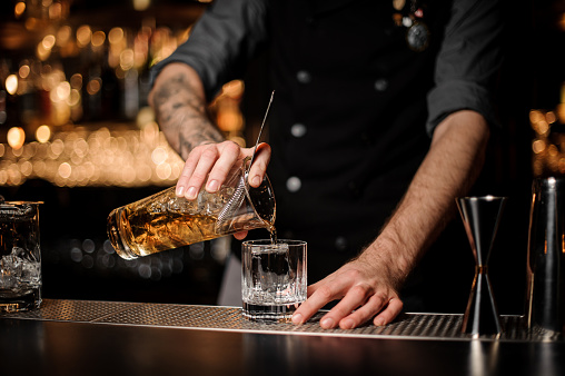 Male bartender with tattoos pours alcohol cocktail adding cold whiskey in glass