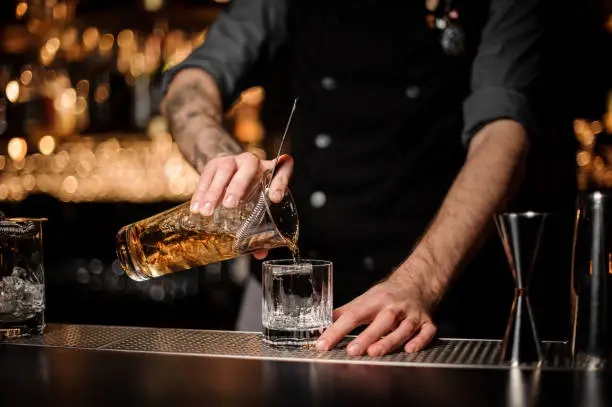 Photo of Bartender pours cocktail adding whiskey in glass