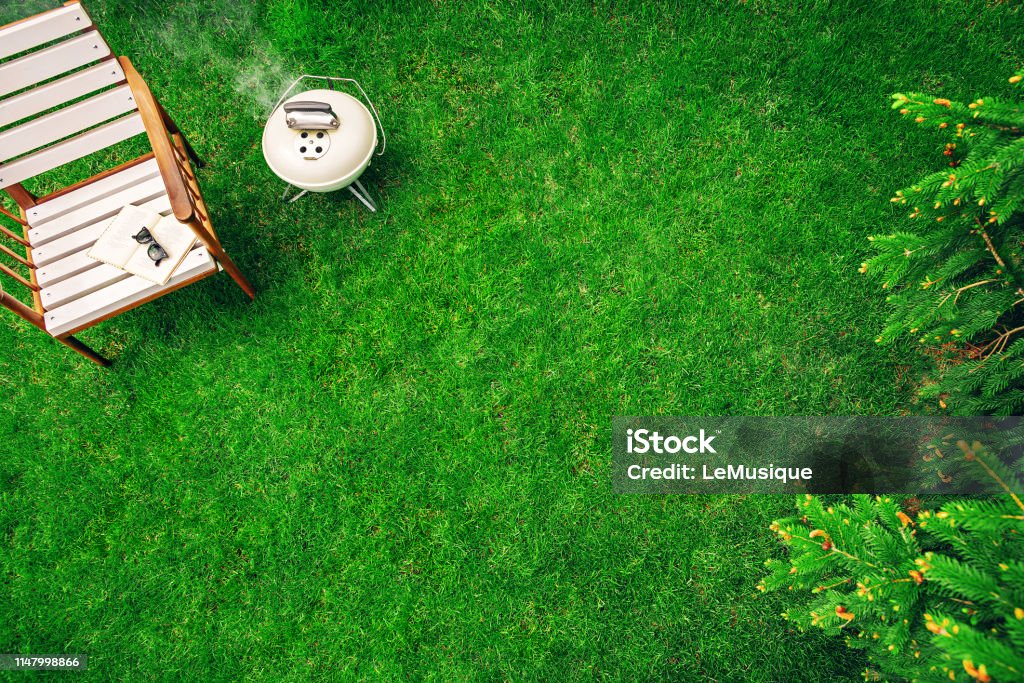 Ivory colored grill on the grass near the wooden armchair with a book and glasses. Top view Barbecue Grill Stock Photo