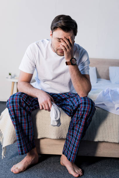 istock handsome sleepy man sitting on bedding and holding white socks 1148004730