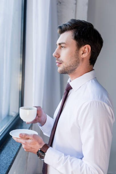 istock handsome serious man with coffee cup standing by window and looking away 1148004762