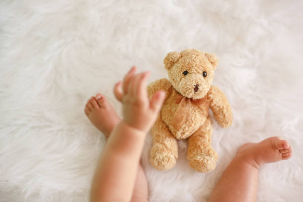 Top view of baby's hand reaching for teddy bear toy. Baby is sitting on fluffy white background.
