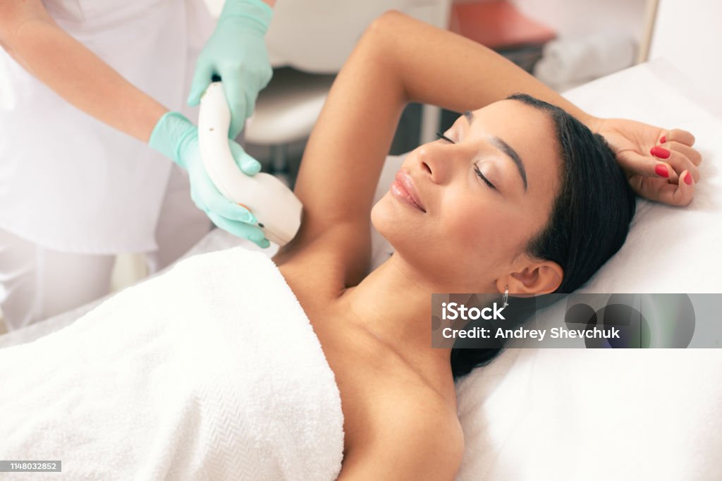 Smiling woman undergoing laser hair removal on her armpit Calm young woman lying with closed eyes and putting on arm up while having laser hair removal procedure on it Laser Stock Photo