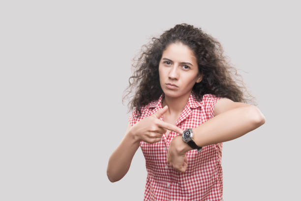 портрет разъяренной молодой женщины, указывающей пальцем на наручные часы на сером фоне - checking the time women impatient wristwatch стоковые фото и изображения