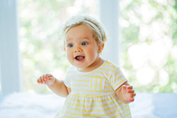 Portrait of nine months old cute baby girl in yellow dress sitting on fluffy white blanket with mouth open, looking excited.